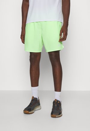 Mann trägt hellgrüne Sportshorts, weiße Kniestrümpfe und schwarz-braune Laufschuhe und steht vor einem weißen Hintergrund.