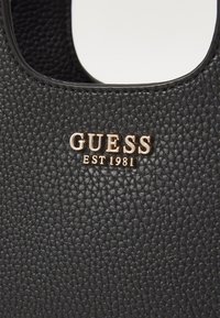 Gros plan sur cuir noir texturé avec logo doré "GUESS EST 1981" sur une découpe de poignée de sac à main.