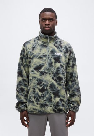 Homme portant un pull polaire tie-dye vert et noir avec une fermeture éclair à l'avant, debout devant un fond clair uni.