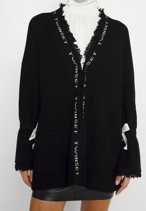 Cardigan noir ouvert à l'avant avec le texte "TWINSET" le long des bords frangés, associé à un chemisier blanc à volants et une jupe en cuir noire.