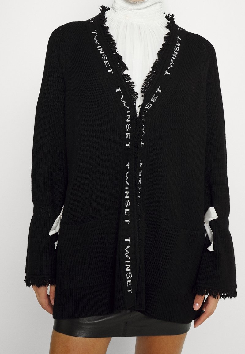 Cardigan noir ouvert à l'avant avec le texte "TWINSET" le long des bords frangés, associé à un chemisier blanc à volants et une jupe en cuir noire.