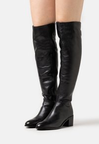 Botas de cuero negro por encima de la rodilla con un acabado elegante, punta afilada y tacón cuadrado. Cuenta con una cremallera lateral para un fácil uso.