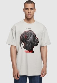 Ung mand med kort hår iført lysegrå T-shirt med sideprofil af en person med flettet hår og rød tekst "Flying High."