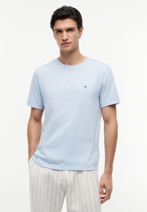 Mann iført lyseblå kortermet t-skjorte og hvite bukser med tynne, vertikale striper, står med én hånd i lomma.