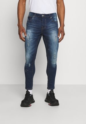 Vaqueros skinny fit de color azul oscuro, con detalles desgastados, un diseño clásico de cinco bolsillos y costuras en contraste. La tela parece ser de denim con un ligero brillo.