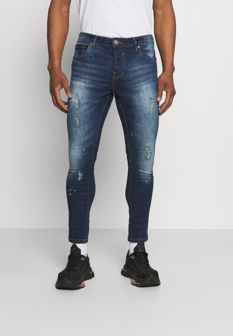 Skinny-fit dark blue jeans, featuring distressed details, a classic five-pocket design, and contrast stitching. Fabric appears denim with slight sheen.