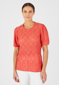 Top in pizzo rosso con maniche a palloncino corte, caratterizzato da un motivo a rombi e dettagli a giorno. Collo a giro e taglio dritto. Indossato con pantaloni bianchi.