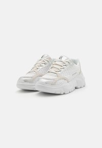 White athletic shoes with mesh and suede accents, featuring a thick sole, textured design, and contrasting laces. Logo on tongue.