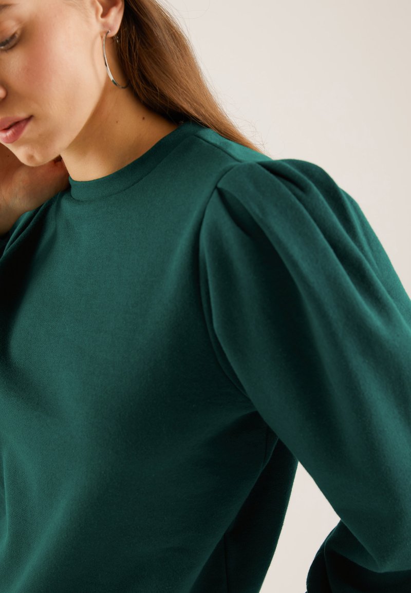 Green sweatshirt with a high neckline and puffed sleeves. Fabric appears soft with a smooth texture. Model shows a close-up view.