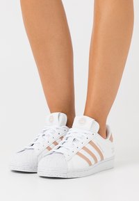 Vita lädersneakers med tre ljusrosa ränder, gummitå, och fläckad vit sula, med varumärke på tungan och sidan.