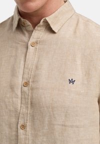 Camicia in lino beige chiaro con chiusura a bottoni, bottoni in legno e logo blu ricamato sul petto. Texture morbida e design casual.