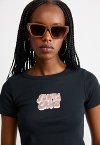 Svart t-skjorte med en fargerik "SANTA CRUZ" grafikk i sentrum. Modellen har på seg lysebrune cat-eye solbriller og har flettet hår.