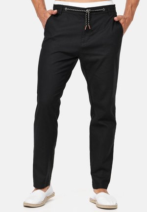 Schwarze Freizeithose mit weißem und schwarzem Kordelzug, getragen mit weißen Slip-On-Schuhen, Hände in den Taschen, auf weißem Hintergrund.