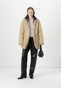 Krémová prošívaná péřová bunda s šedým kožešinovým límcem, černé kožené kalhoty a boty s špičatou špičkou. Drží malou černou kabelku.