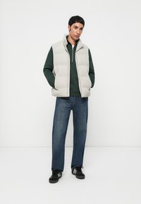 Lys grå puffer vest over en mørkegrøn langærmet skjorte, parret med blå jeans og sorte sneakers. Enkel design, lynlåslukning foran.