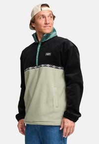 Pullover jacket with a black corduroy upper and green fleece lower, features a turquoise mock neck, geometric trim, and front pockets.