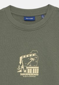 T-shirt in cotone verde oliva con colletto rotondo e stampa grafica che raffigura una scena di mercato in beige, con la scritta "SUPERMERCADO" e "Accetta nessuna sostituzione".