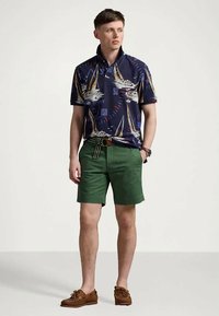 Νεαρός άνδρας φορώντας ναυτικό πόλο με σχέδια ιστιοφόρων, πράσινα σορτς με ριγέ ζώνη και καφέ σκαφάκια, που στέκεται μπροστά σε απλό φόντο.