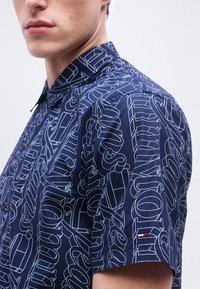 Νέος άνδρας που φοράει μπλε σκουρό μπλουζάκι με κοντά μανίκια, κουμπωτό, με άσπρα αφηρημένα γραφικά κείμενα και ένα μικρό κέντημα λογότυπο σε κόκκινο, άσπρο και μπλε χρώμα.