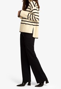 Pull rayé crème et noir avec texture côtelée, fentes sur les côtés et manches ajustées, associé à un pantalon large noir et des escarpins pointus.