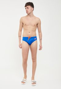 Slip da bagno blu con elastico in vita e cordino arancione. Presenta un piccolo logo su un lato. Indossati con sandali bianchi.