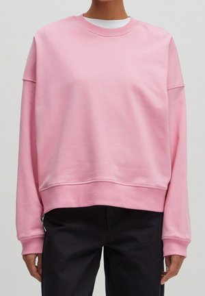 Person trägt ein locker sitzendes, hellrosafarbenes Sweatshirt mit Rundhalsausschnitt über einem weißen Hemd und schwarze Hose, stehend vor einem schlichten Hintergrund.
