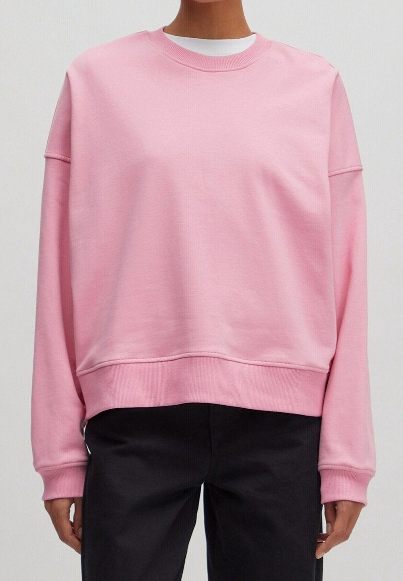 Personne portant un sweat-shirt rose clair à col rond, ample, sur une chemise blanche avec un pantalon noir, debout devant un fond uni.
