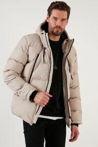 Giacca padded beige con cappuccio, caratterizzata da dettagli neri, tasche con zip e una texture morbida. Indossata sopra un maglione nero e jeans scuri.