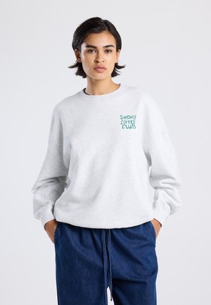 Persoon met kort donker haar draagt een lichtgrijze sweatshirt met de tekst "Sunday Coffee Club" en losse blauwe broek, met de handen in de zakken.