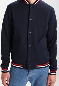 Marineblauwe fleece varsityjack met een geribbelde kraag met rode en witte strepen, voorkant met drukknoopsluiting en zijzakken.