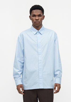 Jeune homme portant une chemise bleu clair à manches longues, debout devant un fond blanc uni.