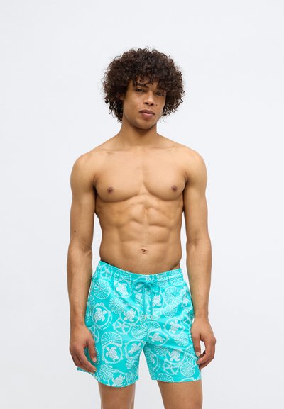 Moški brez majice s kodrasto pričesko, oblečen v turkizne kopalne hlače, okrašene z belimi morski skvočki in školjkami, na enobarvni svetli podlagi.