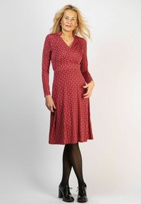 Punaisen värinen mekko, jossa on pitkät hihat, kietaisusuunnittelu, valkoinen abstrakti kuvio, polven pituinen helma, yhdistetty mustiin nauhallisiin kenkiin ja sukkahousuihin.