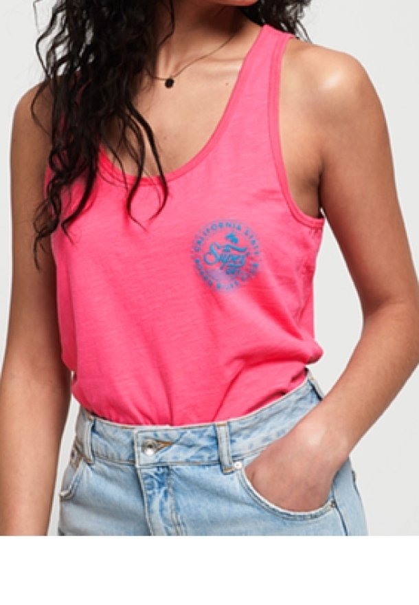 Kobieta nosząca jasnoróżowy top z cienkimi ramiączkami z małym niebieskim nadrukiem na piersi, włożony do jasnoniebieskich, wysokich spodni jeansowych.