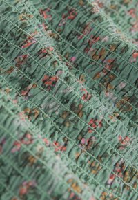 Green floral-patterned fabric with gathered texture. Small pink, orange, and brown accents on textured surface. Soft, stretchy cotton material.