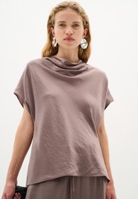Top corto a maniche corte di un delicato colore viola chiaro, con scollatura drappeggiata, realizzato in un morbido tessuto testurizzato. Indossato con grosse orecchini in tono argento.