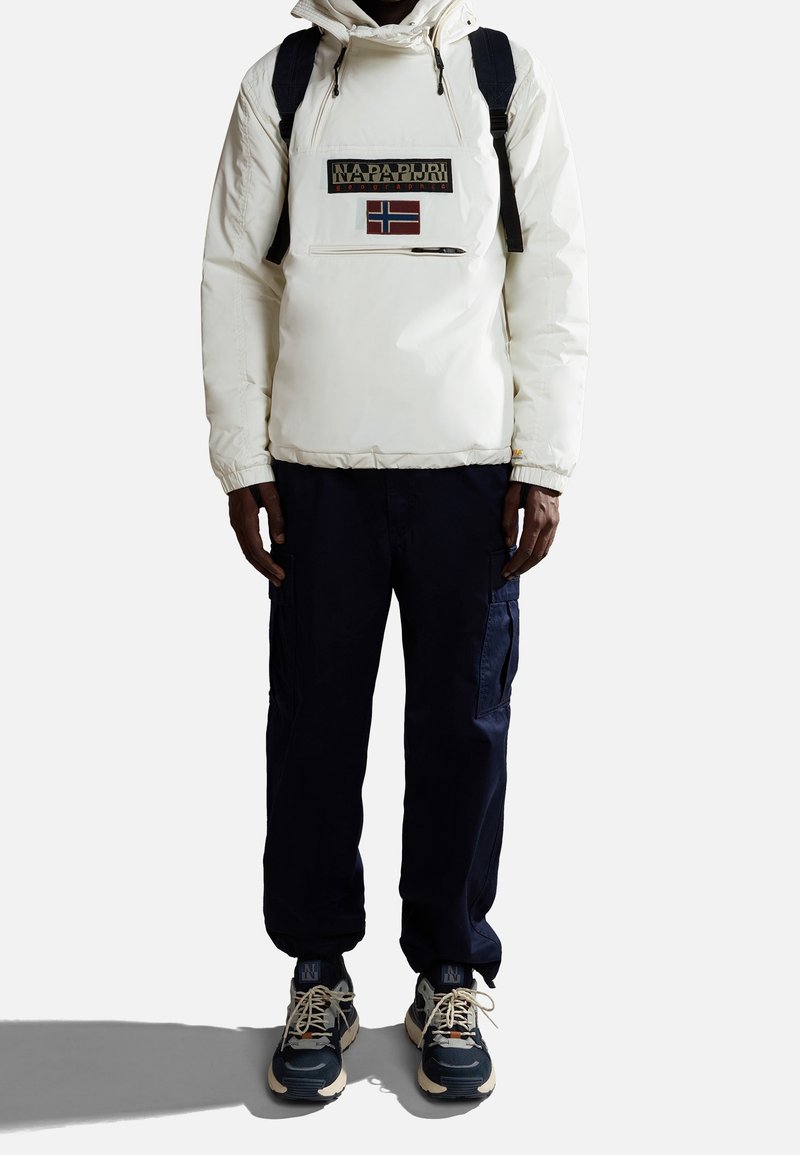Personne portant une veste blanche Napapijri avec un patch du drapeau norvégien, un sac à dos noir, un pantalon cargo foncé et des baskets, debout devant un fond blanc.
