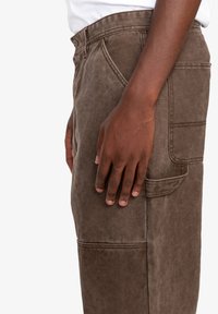 Brown cotton cargo pants with a side pocket, textured finish, and visible stitching details, featured on a model's side view.