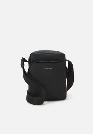 Petit sac à bandoulière noir compact avec poche avant zippée et bandoulière réglable, arborant un petit logo "Calvin Klein" sur le panneau avant.