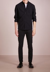 Černá dlouhá košile s límečkem na knoflíky a přední kapsou, kombinovaná s černými slim-fit džíny s dírami na kolenou a černými šněrovacími botami.