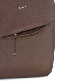 Bolsa de tela marrón con un logo de Nike plateado, un bolsillo con cremallera y una superficie suave y texturada. Diseño sencillo con acentos funcionales.