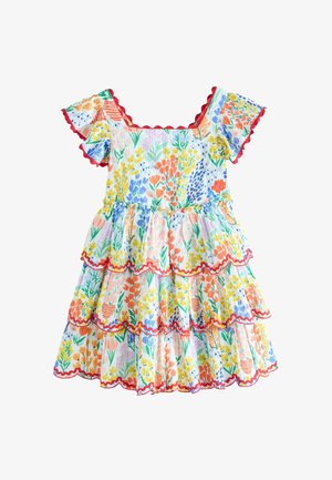 Junges Mädchen, das ein buntes Blumenkleid mit Rüschensaum und passenden Sandalen trägt, vor einem schlichten Hintergrund stehend.