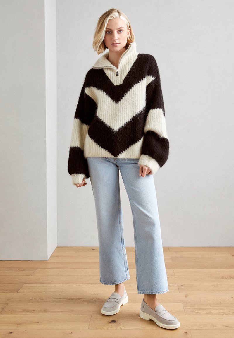 Suéter de gran tamaño con patrón de chevron en negro y crema, con cuello zip y textura acanalada. Combinado con vaqueros de pierna ancha en azul claro y mocasines grises.