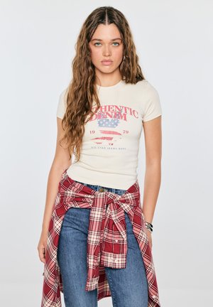 Młoda kobieta z długimi kręconymi włosami nosi kremowy t-shirt z nadrukiem, niebieskie dżinsy oraz czerwoną koszulę w kratę zawiązaną wokół talii, stoi na jednolitym tle.