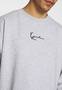 Gros plan d'une personne portant un sweat-shirt gris clair à col rond avec un petit logo noir brodé sur la poitrine.