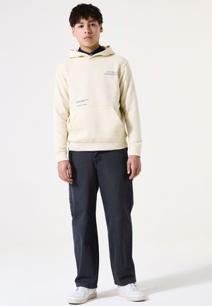 Sweatshirt à capuche crème avec poche frontale, orné de texte bleu sur la poitrine. Associé à un jean sombre et des baskets blanches.