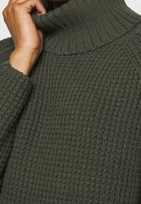Tloušťkový pletený svetr s rolákem v tmavě zelené barvě. Má žebrovanou texturu, široký límec a design se spadenými rameny.
