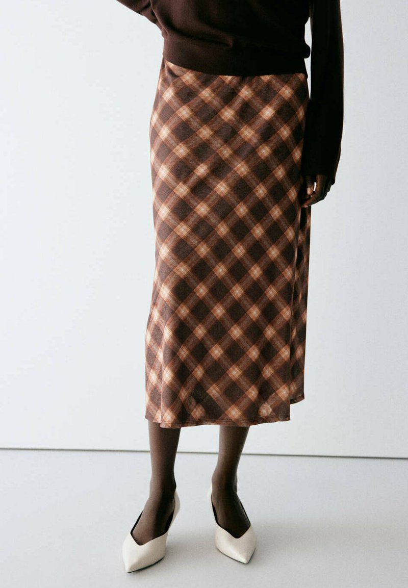 Femme portant une jupe midi marron à carreaux, des collants noirs, un haut à manches longues foncé et des chaussures blanches à bouts pointus, debout devant un fond uni.