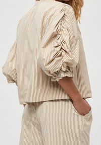 Beige gestreifte Bluse mit puffigen Ärmeln, gerafften Bündchen und lockerer Passform. Der Stoff wirkt glatt mit vertikalen Streifen.