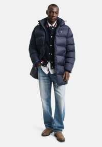 Marineblå pufferjakke med hætte, med lynlås lukning og store baffles. Båret over en marineblå sweater og en stribet skjorte, parret med lyse jeans og støvler.